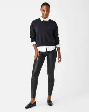 A woman stands confidently against a plain white background, wearing the SPANX Aireessentials Crew Neck Pullover and matching sweatpants made from luxurious comfort spacer fabric. She has her hair pulled back and her left hand is raised slightly. She smiles gently with her lips closed.