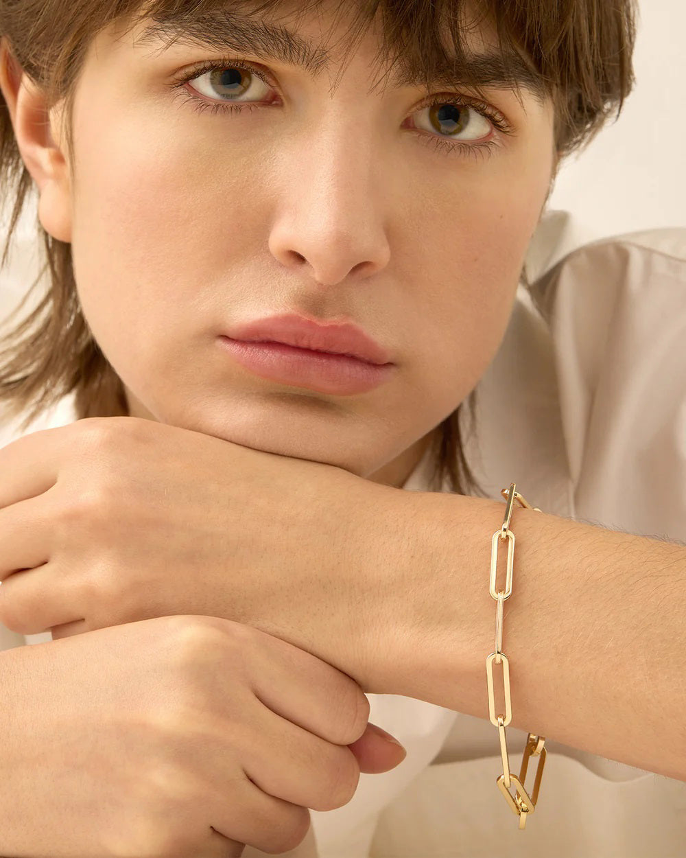 A person with medium-length hair wearing a white shirt is gazing ahead, resting their chin on crossed arms. Their wrist is adorned with the JENNY BIRD - ANDI SLIM BRACELET, its high-polish gold finish gently catching the light. The background is softly blurred.