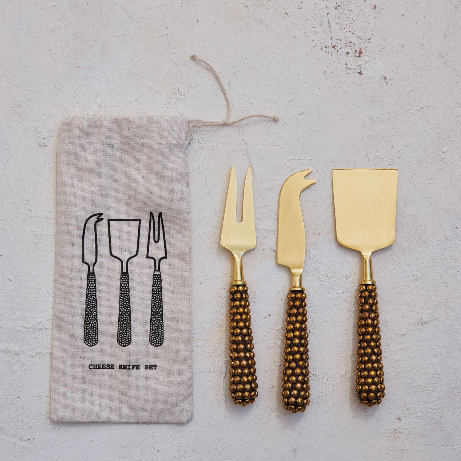 A trio of gold stainless steel cheese knives by Creative Coop, each featuring a beaded handle, is elegantly showcased beside a fabric pouch. The pouch is adorned with black illustrations of the knives and the phrase "Cheese Knife Set." All elements rest on a textured white surface, highlighting their elegance and eco-friendly packaging.