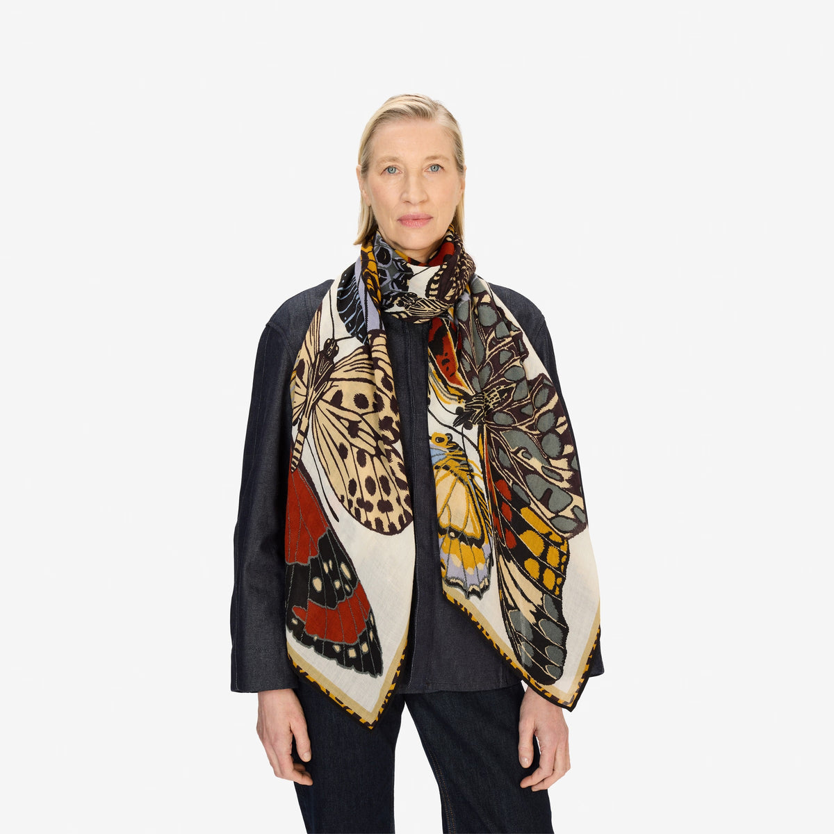 A person dressed in dark attire models the INOUI EDITIONS PAPILLONS (BUTTERFLIES) wool scarf in white by INOUI, featuring a colorful butterfly wing pattern, against a plain white background.