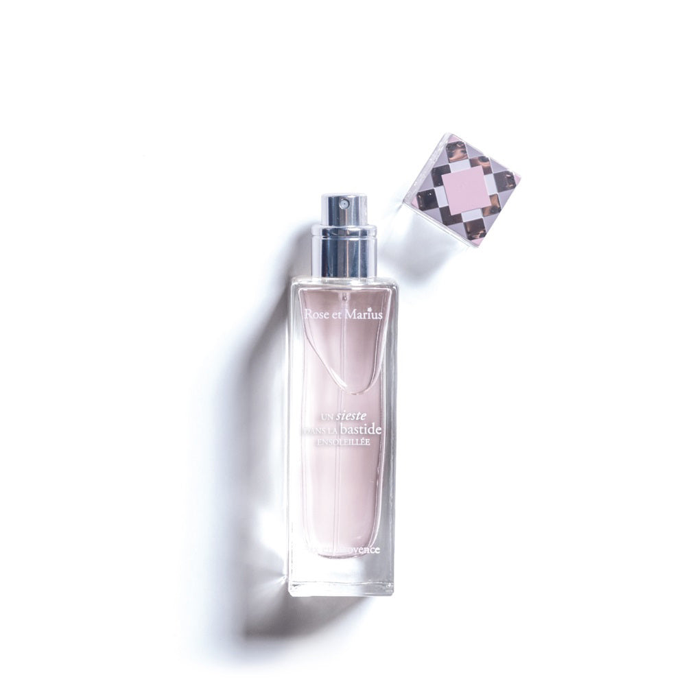 A rectangular glass perfume bottle with a silver spray nozzle and square cap beside it casts a shadow on white. The label reads &quot;ROSE ET MARIUS - A SIESTA IN A SUNLIT HOME EAU DE PARFUM 30ML,&quot; evoking the scent of Provence.