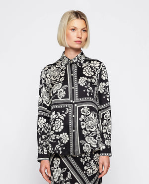 A woman with blonde, chin-length hair is wearing the MIRTO 1956 - BLACK AND WHITE PRINT FLUID BLOUSE. Embracing the intricate flower squares patchwork print of MIRTO fashion, she stands neutrally against a plain white background.