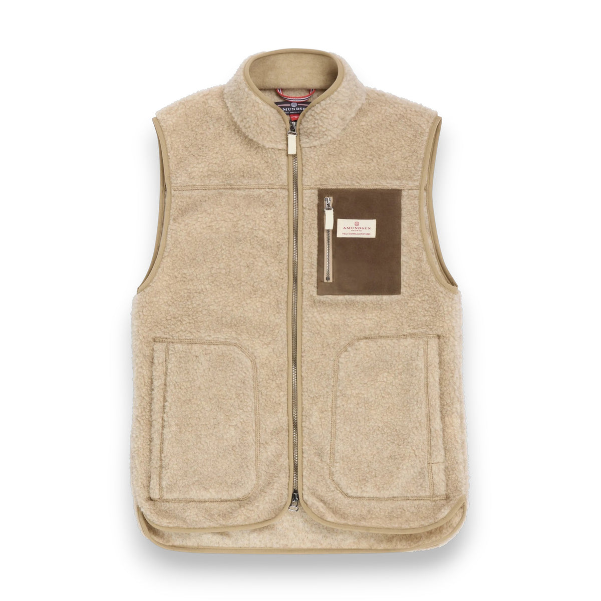 A man stands on a patterned rug wearing the AMUNDSEN SPORTS - HEROES FLEECE VEST NATURAL over a green ribbed sweater, beige pants, brown boots with green ankle fabric, and holding a green duffel bag. The background is plain and neutral.