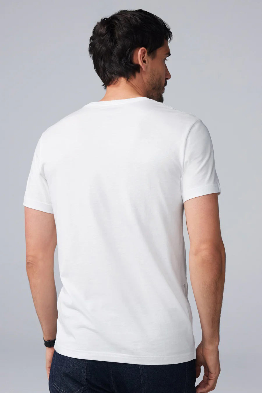 A man with short dark hair and a beard wears the MACK WELDON SILVER CREW NECK T-SHIRT IN WHITE and dark pants, standing in profile with his head slightly bowed and one hand in his pocket against a light gray background.