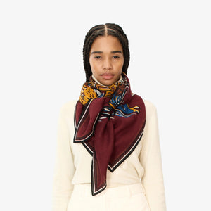 A person is wearing a cream-colored sweater with the INOUI EDITIONS - WESTERN SQUARE SCARF from INOUI, a large, burgundy wool scarf adorned with unique prints draped around their neck. The image focuses on the upper body, showcasing the intricate design of the scarf against a plain white background.