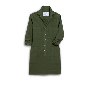 A woman with long brown hair smiles indoors, wearing the FRANK & EILEEN SOUTHERN IRELAND UTILITY DRESS in Hunter Green. She stands by a beige armchair and lamp, with patterned wallpaper in the background.