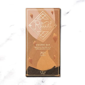 The RITUAL CHOCOLATE - SMORES CHOCOLATE BAR 70% features a rectangular shape adorned with a speckled golden topping, reminiscent of marshmallow flavors, set against a white marble background. The bar has a slightly textured surface due to the glittery gold flecks covering it.