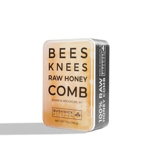 A rectangular tin container labeled "BUSHWICK KITCHEN - 'BEES KNEES' RAW HONEYCOMB" by Bushwick Kitchen. The off-white tin with black and orange text states "Born in Brooklyn, NY" and "Net Wt 7oz (198g)." One side is marked "100% Raw Honeycomb," containing pure, edible honey comb.