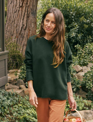 Close-up image of a person standing outdoors, wearing the FRANK & EILEEN Anna Long Sleeve Capelet in Evergreen with cozy sleeves and orange pants. Only part of their upper body and arms are visible. The scene is set against a blurred background of greenery.