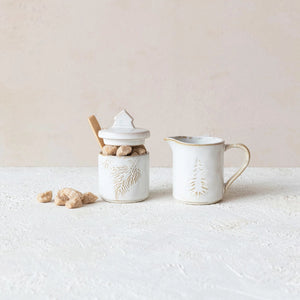 Beside a matching creamer jug, you'll find the STONEWARE SUGAR POT WITH SPOON by CREATIVE COOP. Both pieces, part of an elegant stoneware collection, feature white ceramic with embossed pine tree designs and include a subtle rustic finish that adds charm to their appearance.