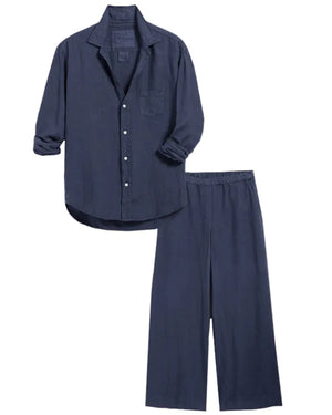 A person wearing a relaxed, navy blue button-up shirt and matching wide-leg silhouette pants from the FRANK & EILEEN Tuscany Getaway Set, including the FRANK AND EILEEN - MAISIE WIDE LEG LINEN PULL ON PANT, with hands in pockets, stands poolside on a sunny day. They carry a beige woven tote bag with blue and red stripes, have short curly hair, and wear sunglasses and sandals.