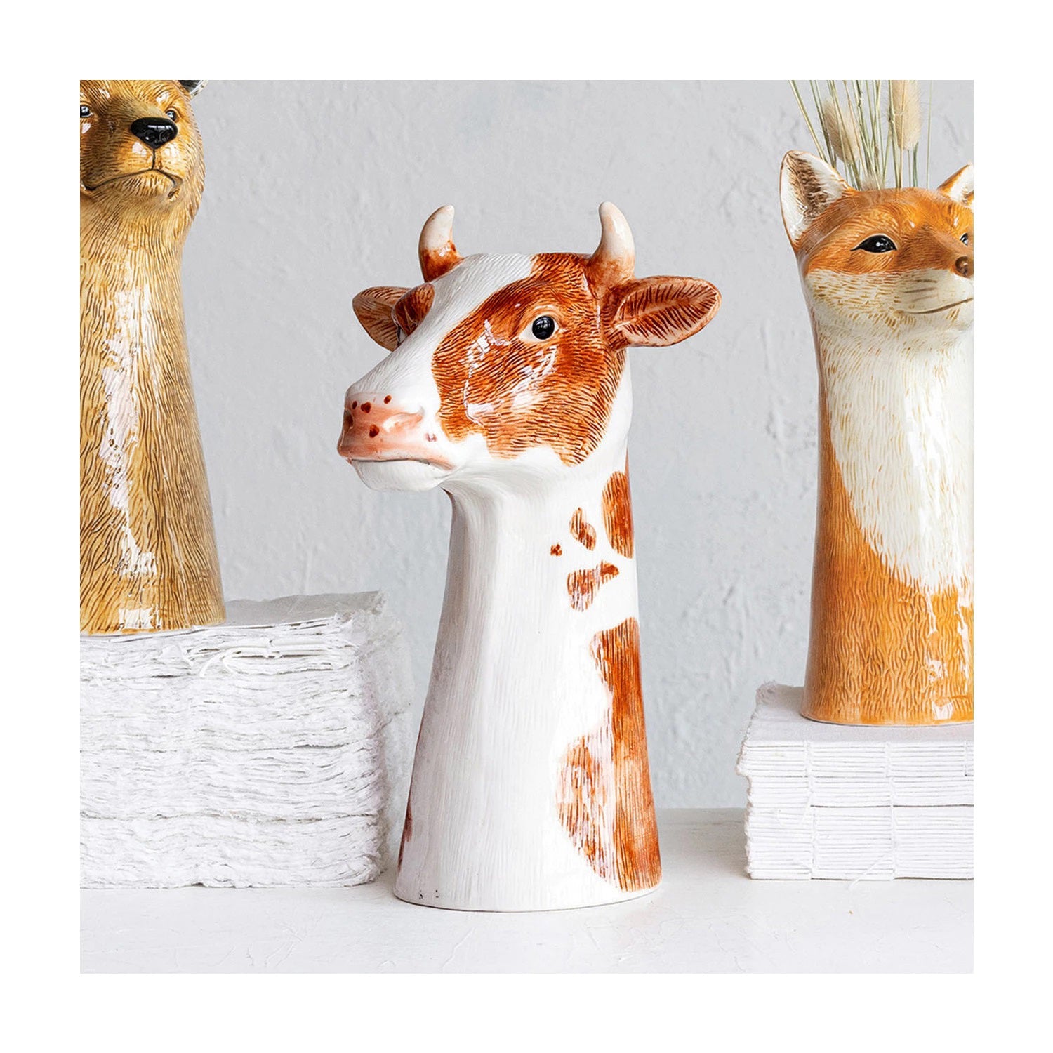 The CREATIVE COOP CERAMIC COW VASE is a hand-painted stoneware vase shaped like a cow's head and neck, featuring a farmhouse style with brown-and-white details, horns, and a realistic expression. Its top opening adds functionality to its captivating design against a plain white background.