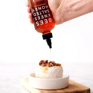 A red bottle of BUSHWICK KITCHEN - 'BEES KNEES' SALTED HONEY is shown. The label reads "BORN IN BROOKLYN, NY" and "NET WT 12.5 OZ (354g)." This gluten-free treat features wildflower honeys perfected with a hint of salt and comes with a black pointed cap.