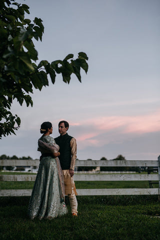 A couple dressed in elegant traditional attire stands closely together outdoors near a white fence at sunset, with a leafy tree branch overhead and a pinkish sky in the background.