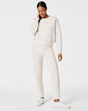 A person stands against a white background, smiling and looking down with one hand touching their hair. They are wearing a matching light beige SPANX AIRESSENTIALS TAPERED PANT and sweatshirt set made from ultra drapey, lightweight spacer fabric, paired with white sneakers.