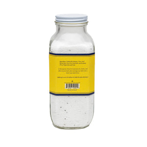 The BAUDELAIRE - BOOST BATH SELTZER 18OZ comes in a clear glass bottle with a yellow label, showcasing white bath salts, essential oils, and visible small herbs inside. The label provides ingredients and usage instructions. The bottle features a screw-on metal cap and is filled nearly to the top.