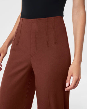 A person stands wearing a black short-sleeve top and the SPANX Stretch Twill Cropped Wide Leg Pant in brown, featuring CoreSure Tech for comfort. Paired with black sandals, they smile brightly, hands relaxed by their sides against a plain light gray background.