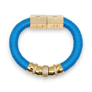 The HOLST + LEE - LUXE CLASSIC BRACELET by HOLST & LEE features an elegant orange cord with gold-plated accents, including a rectangular gold clasp adorned with several gold and diamond-encrusted beads near the clasp.