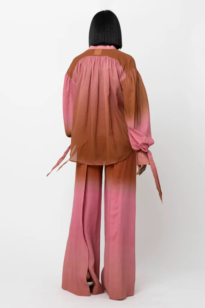 A person stands confidently wearing a stylish outfit consisting of the HOPE FOR FLOWERS Wrist Tie Blouse and matching pants in a gradient pink to brown color. Made from sustainable fabric, the blouse features balloon sleeves and a relaxed collar. The person has a serious expression and shoulder-length straight hair.