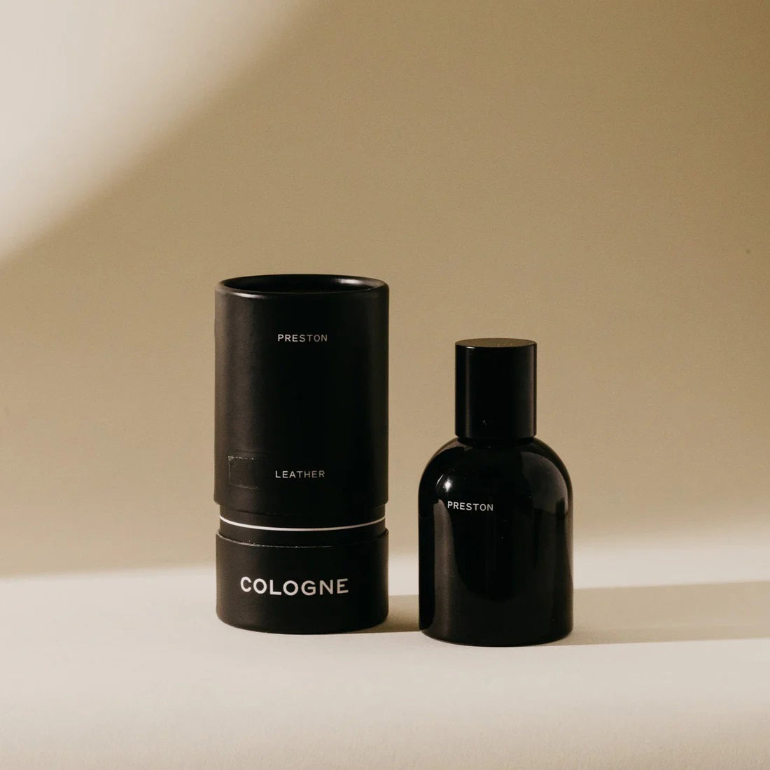 A sleek black bottle of PRESTON - COLOGNE 1.7OZ stands next to its matching cylindrical case. Both items are set against a neutral beige background, creating a minimalist and sophisticated look. The black packaging, made from biodegradable materials, features simple white text labeling the product with the brand name PRESTON.