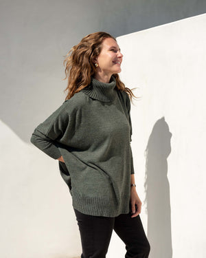 A woman with wavy hair is smiling and looking to the right, wearing a cozy green MERSEA - CATALINA TURTLENECK SWEATER and black pants. She stands against a white wall, with sunlight casting a shadow behind her.