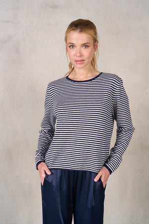 A person with blonde hair in a ponytail is shown from the back, wearing the ROSE & CROWN STRIPE CASHMERE CREWNECK SWEATER and dark blue trousers. The outfit exudes a sophisticated look against the plain, light-colored wall backdrop.