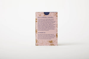 A rectangular pink box with brown floral patterns contains JACOBSEN SALT - SALTY HAZELNUT CARAMELS BOX by JACOBSEN SALT CO. The back of the box features descriptive text about the product's ingredients and origin, highlighting the use of Oregon sea salt and Jefferson hazelnuts for a rich flavor.