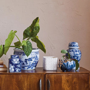 A wooden cabinet with silver handles is topped with several ceramic vases. Two vases feature blue and white patterns, one holding a green leafy plant. A white square vase and the CREATIVE COOP SQUARE PLANTER WITH DEBOSSED WOVEN PATTERN, each with another plant, complete the charming display.