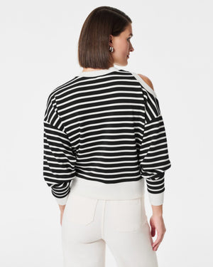 A person with short hair poses against a white background, wearing the SPANX - AIRESSENTIALS COLD SHOULDER TOP in black and white stripes crafted from luxuriously soft material, paired with light-colored pants. Their left hand is placed on their head while their right arm rests at their side.