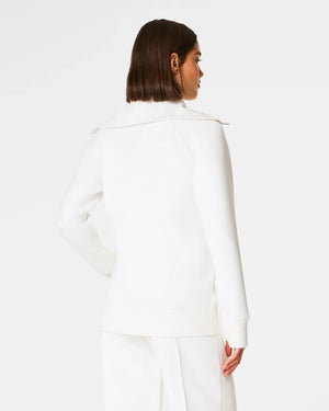 Someone with shoulder-length hair wears a white SPANX AirEssentials Luxe Full Zip Tunic Jacket with matching pants, hands in the tunic's pockets. The plain white backdrop highlights the minimalist loungewear aesthetic.