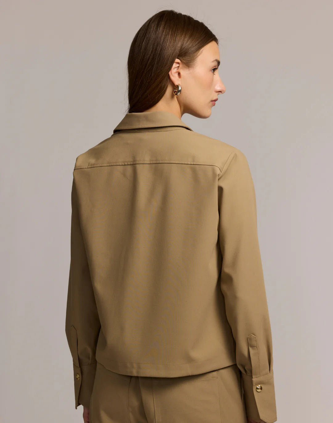 A woman with long brown hair wears the HINSON WU - ASTRID LONG SLEEVE TRAVELER&#39;S TWILL SHIRT JACKET over a black top and tan pants. She stands against a light background, looking confidently at the camera.