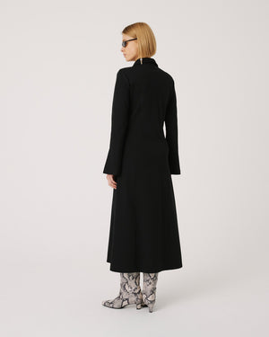 A person with short blond hair stands facing away, wearing a long black coat and snakeskin textured high-heeled boots. The individual, clad in dark sunglasses and the SFIZIO Ponte Knit Midi Dress with long sleeves and a split neck collar, is set against a plain light gray background.