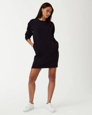 A woman is wearing the SPANX AirEssentials Crew Neck Dress, a black, long-sleeve, knee-length dress from the SPANX brand, with her hands in the functional pockets. She is looking to the side and has long, straight hair. Paired with white sneakers, she stands against a plain white background in this light-as-air fabric attire.