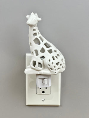 The GIRAFFE NIGHTLIGHT by LILY'S LIGHTS plugs into a wall outlet, featuring a hollow ceramic design with geometric cutouts that allow light to shine through. Its minimalist and decorative look, especially against white-painted walls and outlets, makes it a unique gift for any occasion.