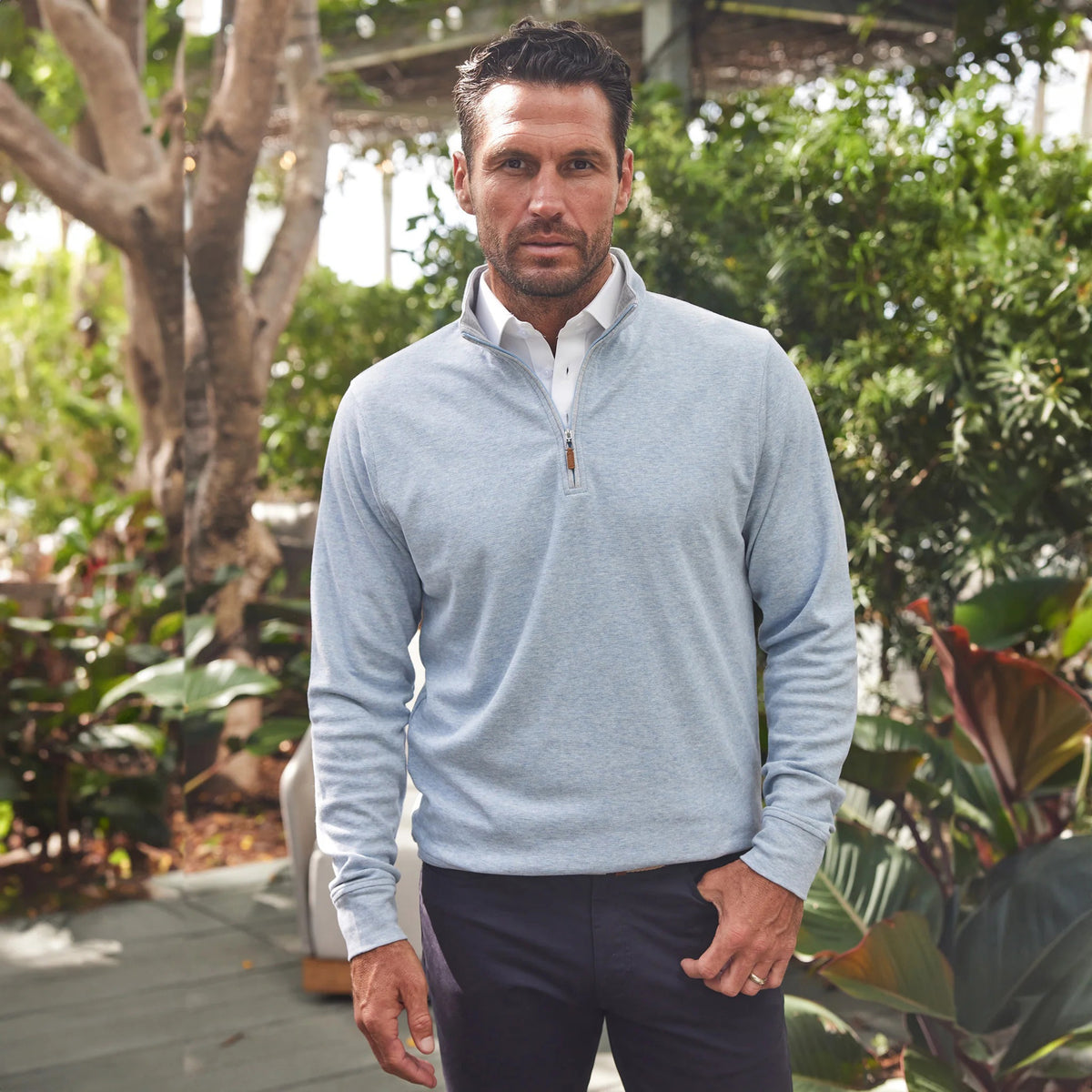 A man smiles outdoors on a sunny day, wearing the COLLARS AND CO Cloud Texture Quarter Zip Pullover in Light Blue over a white polo shirt, with greenery and a shaded path behind him.