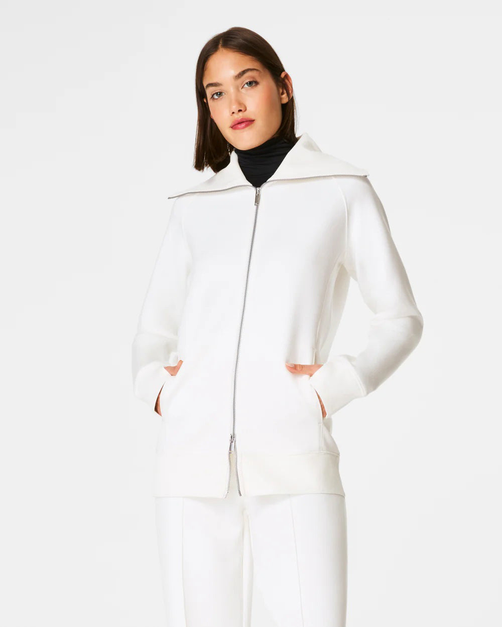 Someone with shoulder-length hair wears a white SPANX AirEssentials Luxe Full Zip Tunic Jacket with matching pants, hands in the tunic's pockets. The plain white backdrop highlights the minimalist loungewear aesthetic.