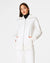 Someone with shoulder-length hair wears a white SPANX AirEssentials Luxe Full Zip Tunic Jacket with matching pants, hands in the tunic's pockets. The plain white backdrop highlights the minimalist loungewear aesthetic.