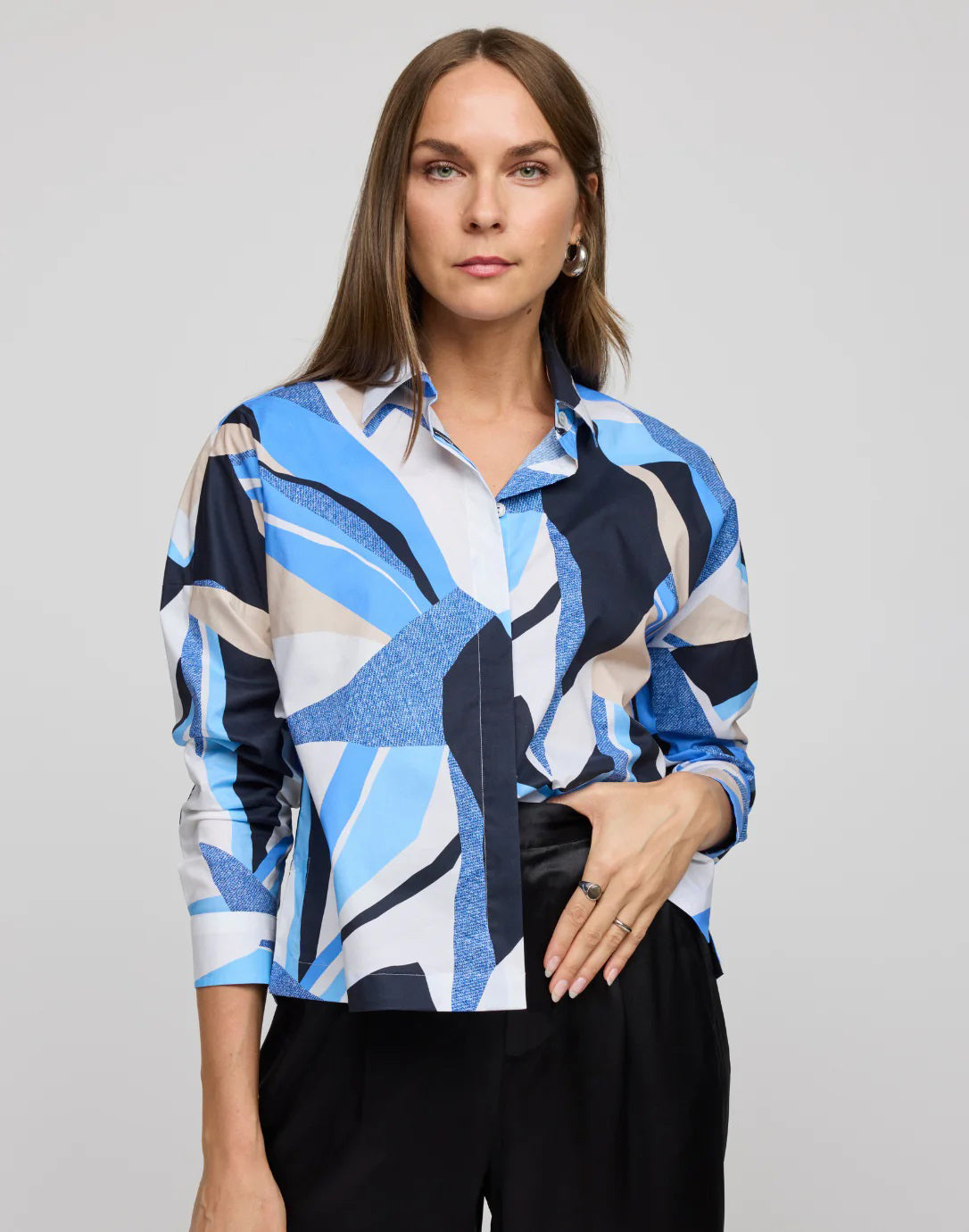 A woman with long brown hair poses against a plain background in the HINSON WU SOPHIE Long Sleeve Abstract Print Shirt in blue, white, and black refined cotton. Her outfit is completed by black pants and a silver earring as her right hand rests on her lower waist.