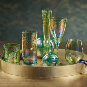 A round, golden tray features a selection of iridescent, textured glassware from the brand ZODAX, each with a capacity of 445 ml. This includes champagne flutes, wine glasses like the APERITIVO RED WINE GLASS - LUSTER GREEN, and tumblers. Two iridescent ornaments are artfully placed among the glassware, set against a dark, blurred background.