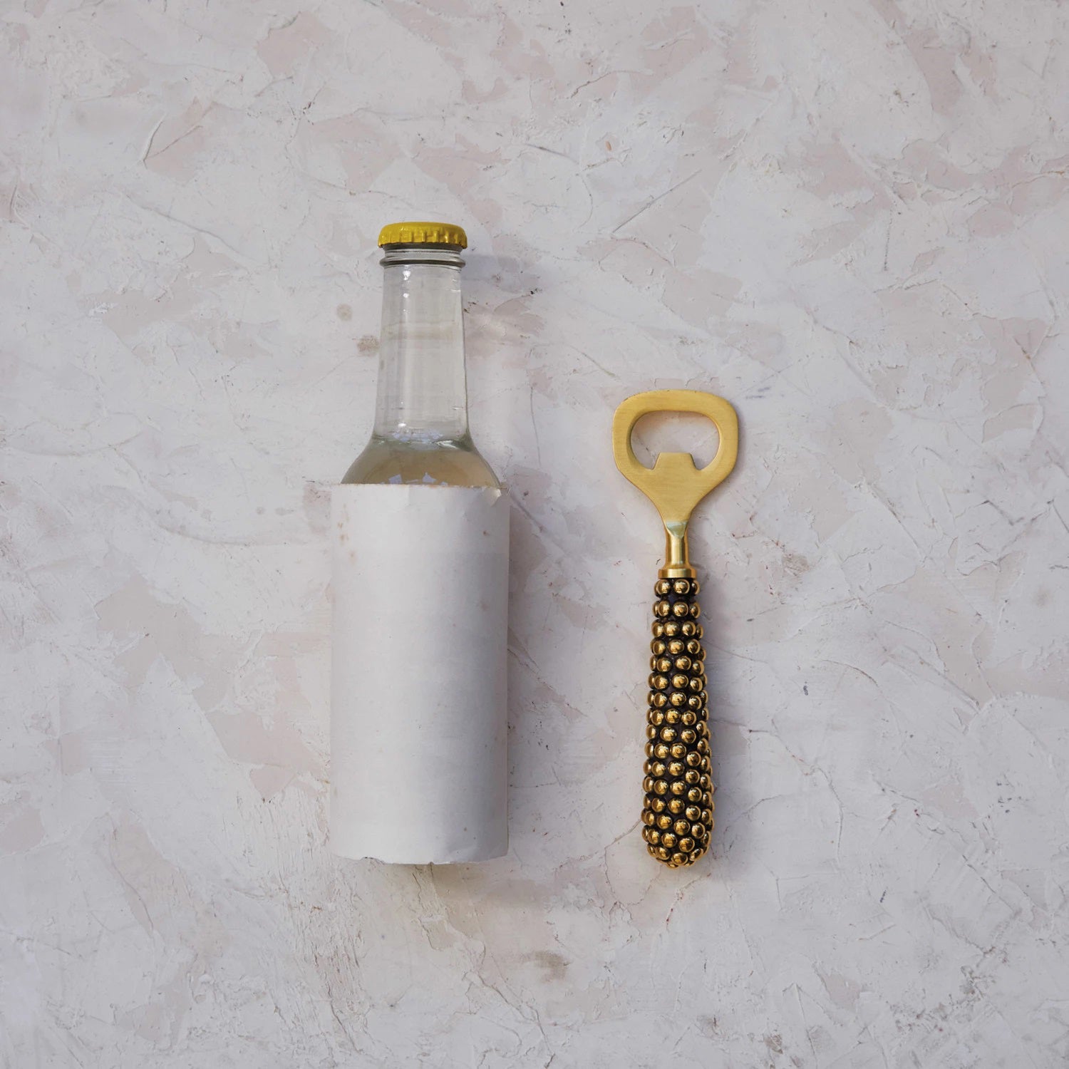 The CREATIVE COOP STAINLESS STEEL BOTTLE OPENER WITH CAST BRASS BEADED HANDLE boasts a golden finish and a corn cob-style design with intricate metallic bumps, offering an elegant nod to classic beaded brass aesthetics while being packaged sustainably for an eco-friendly touch.