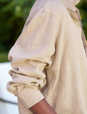 A woman stands on a brick walkway surrounded by greenery and purple flowers. She wears the Tuscany Getaway set, featuring a beige, loose-fitting Eileen Relaxed Button Up Shirt in Linen from FRANK & EILEEN with long sleeves and Maisie pull-on pants, along with brown sandals. She holds a straw hat by her side and smiles slightly, with her hair in loose curls.