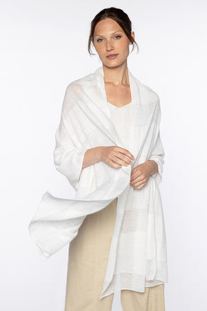 A person with light skin and brown hair is posing in a white, 100% linen outfit with the KINROSS CASHMERE - SHADOW STRIPE LINEN WRAP draped elegantly around their shoulders. They are looking at the camera with a neutral expression against a plain white background.