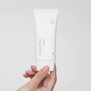 A hand is holding a white tube of MIXSOON CENTELLA SUNSCREEN 50SPF by KOSMIC KBEAUTY SKINCARE. The packaging features minimalist text and design, perfect for delicate skin. The background is solid and light-colored, emphasizing the non-sticky finish of this exceptional product.