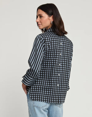 A woman with shoulder-length dark hair wears the HINSON WU - AILEEN 3/4 SLEEVE STRIPE GINGHAM COMBO SHIRT, a black and beige striped blouse featuring a small collar and a partial zip at the front. She pairs it with black pants and stands against a plain white background, one hand in her pocket.