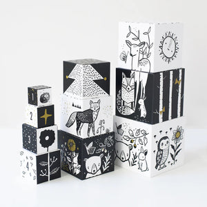 The WEE GALLERY - WOODLAND NUMBERS NESTING BLOCKS by WEE GALLERY are stacked black-and-white nesting blocks adorned with various illustrations of woodland animals, plants, and shapes. The designs include birds, deer, trees, and the sun, with some blocks incorporating gold color accents. These Nesting Blocks help develop motor skills and feature numbers to make counting fun.