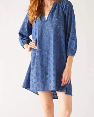 A person is seen wearing the MERSEA DAISY EYELET DRESS by MER SEA, a loose-fitting, knee-length, long-sleeved blue dress adorned with a daisy eyelet cutout pattern. The coastal elegance design features a V-neckline that reveals visible white fabric underneath. Made of 100% cotton, the person has one hand on their hip and the other hanging at their side.