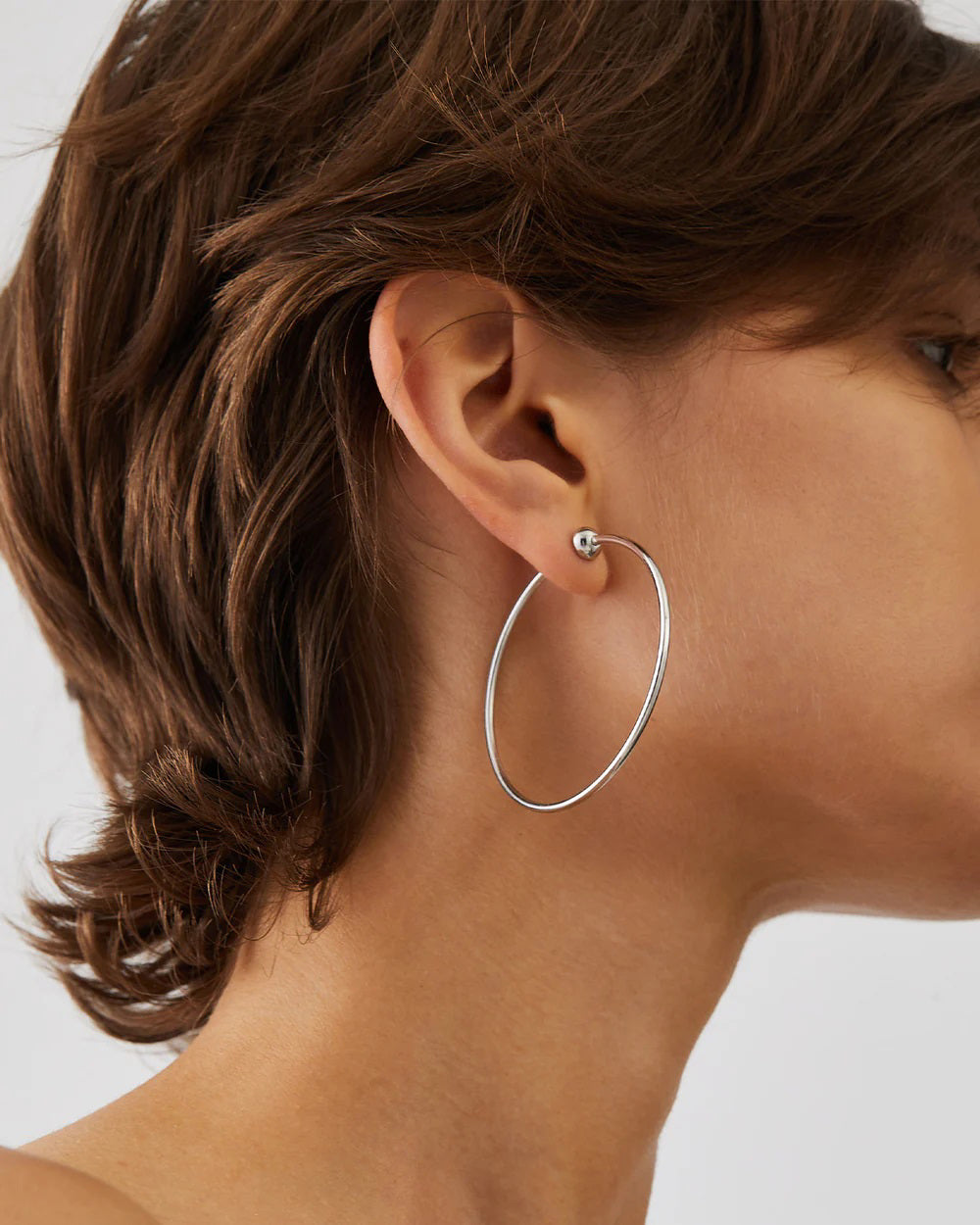 The JENNY BIRD - MEDIUM ICON HOOP EARRINGS, made from high polish stainless steel, feature two thin silver hoops with small beads at the clasp. Set against an off-white background, one earring is upright while the other lies flat.