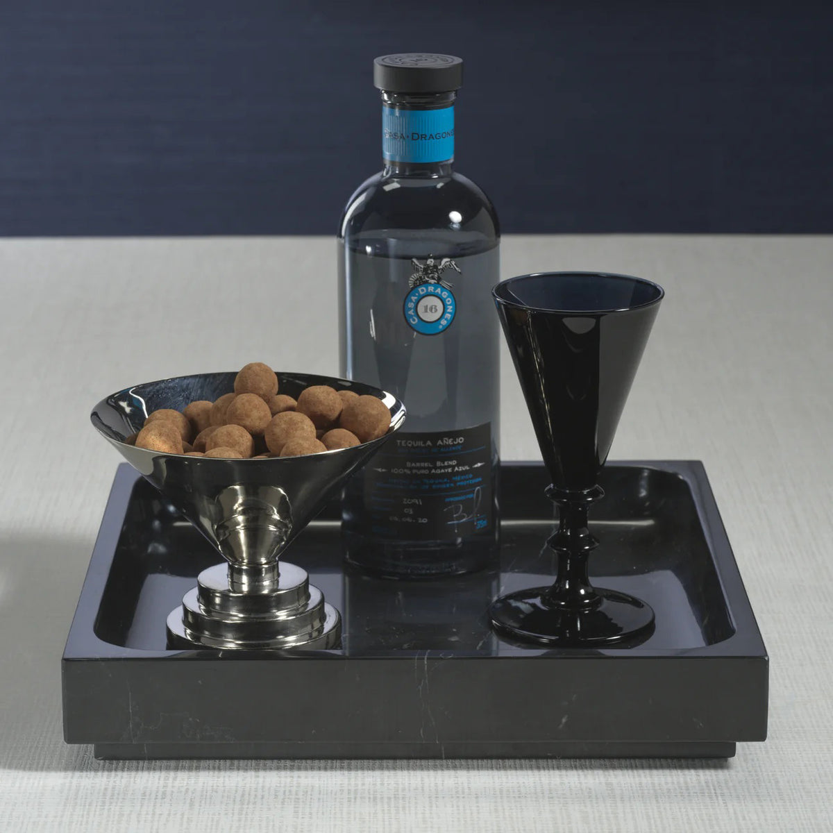 A black bottle of tequila sits on a ZODAX NERO MARQUINA NATURAL MARBLE TRAY - LARGE, accompanied by a black goblet and bowl of chocolate treats, all arranged on a light surface with a dark backdrop.