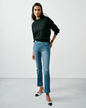 A person stands against a plain backdrop, confidently showcasing their style in SPANX's KICK FLARE JEAN, made with 4-way stretch denim. They pair it with a black sweater and black-and-white heels, their long dark hair cascading as they pose with one hand in their pocket.