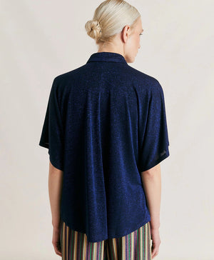 A woman with platinum blonde hair stands with a neutral expression, wearing the MOMONI - BROOKLYN LUREX JERSEY BLOUSE and colorful beaded necklaces. She has her hands in the pockets of her striped trousers with oversize lines. The background is plain and softly lit.